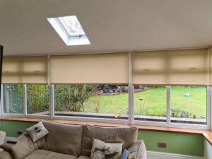 Beige roller blinds partially closed over conservatory windows, with a brown sofa in the foreground and a garden visible outside.