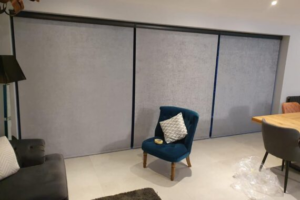 Grey motorised blinds covering a three-panel patio door, with a blue chair on a cream carpet in the foreground, a dark grey sofa to the left, and the end of a wooden table to the right.