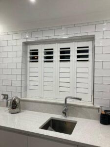 White shutters closed above a kitchen sink and white countertop, with the surrounding wall tiled in white.