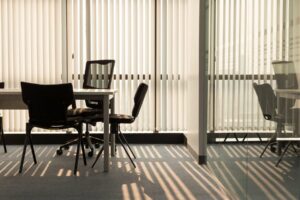 Black office chairs around a white desk, with windows covered by vertical blinds and the background, all reflected in a glass partition to the right.