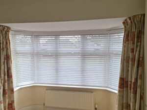 White wooden Venetian blinds closed over a bay window with a radiator below and cream and red patterned curtains at each side.