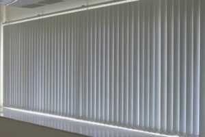 White vertical blinds closed over a wide window, with some light visible at the edges.