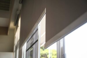 Two grey roller blinds half closed over windows with a green hedge visible through the window.