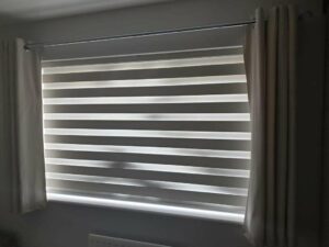 A closed day and night blind in white and grey with cream curtains drawn back at either side.