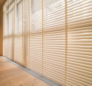 A very broad window covered in extra-wide wooden Venetian blinds in a light cream colour above a wood-effect floor.