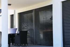 Black Venetian blinds closed over a large patio window, with a white column visible to the left alongside a black chair.