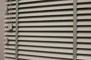 White wooden Venetian blinds partially vented with grey tapes running top to bottom at each side of the frame.