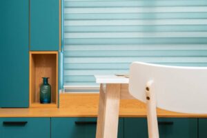 A wooden desk and a white chair behind a set of light blue pleated blinds which are also called concertina blinds