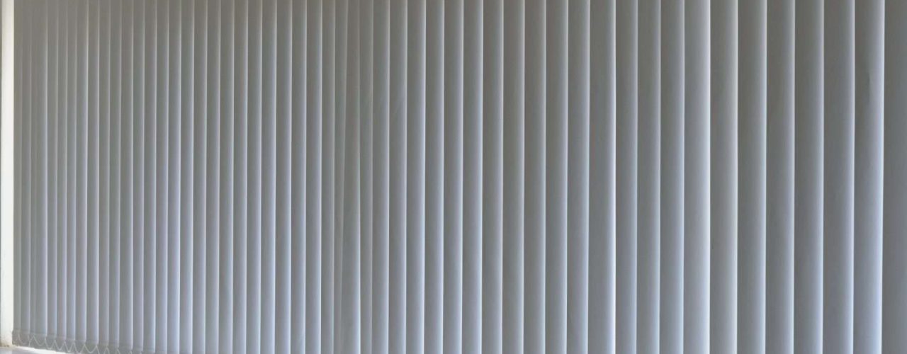 White vertical blinds closed over a wide window, with some light visible at the edges.