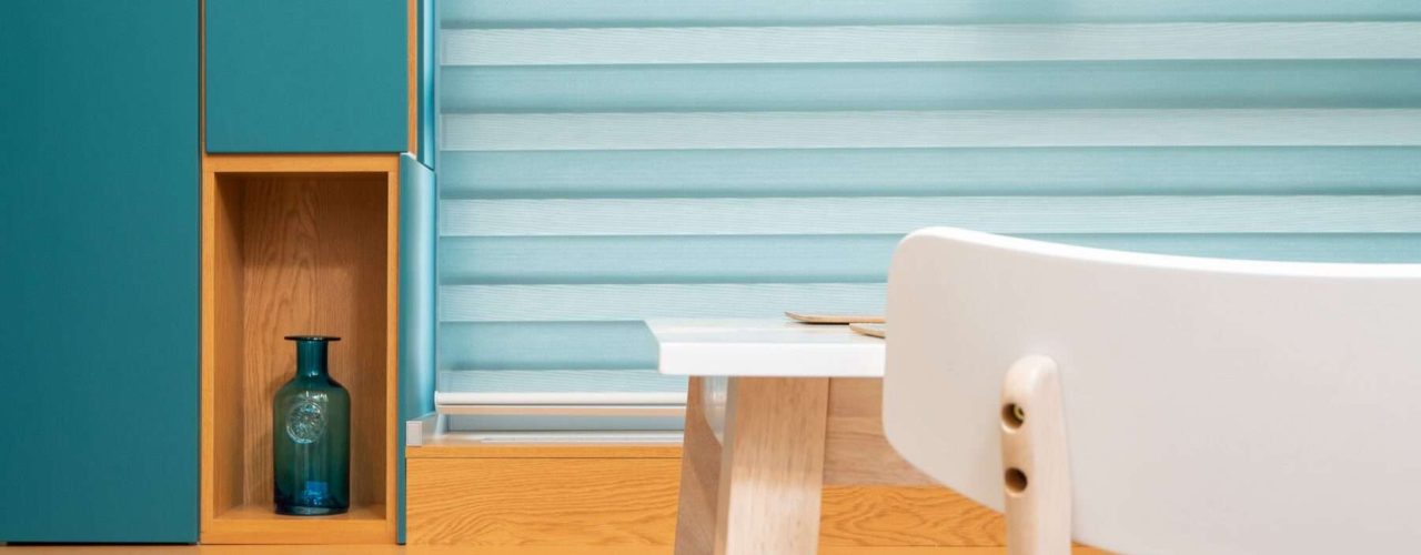 A wooden desk and a white chair behind a set of light blue pleated blinds which are also called concertina blinds