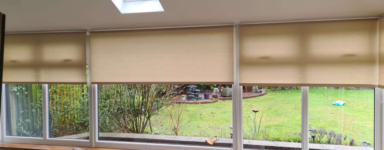 Beige roller blinds partially closed over conservatory windows, with a brown sofa in the foreground and a garden visible outside.