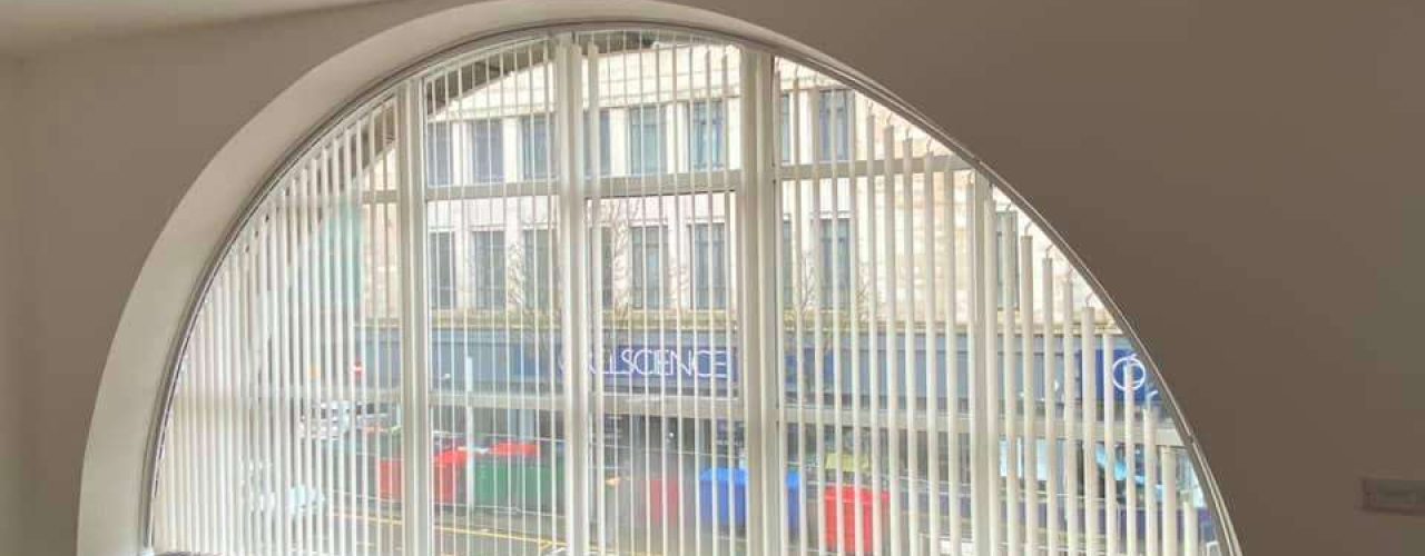 A floor-to-ceiling arch window covered by vertical blinds, with a wooden floor below and a shop visible outside the window.