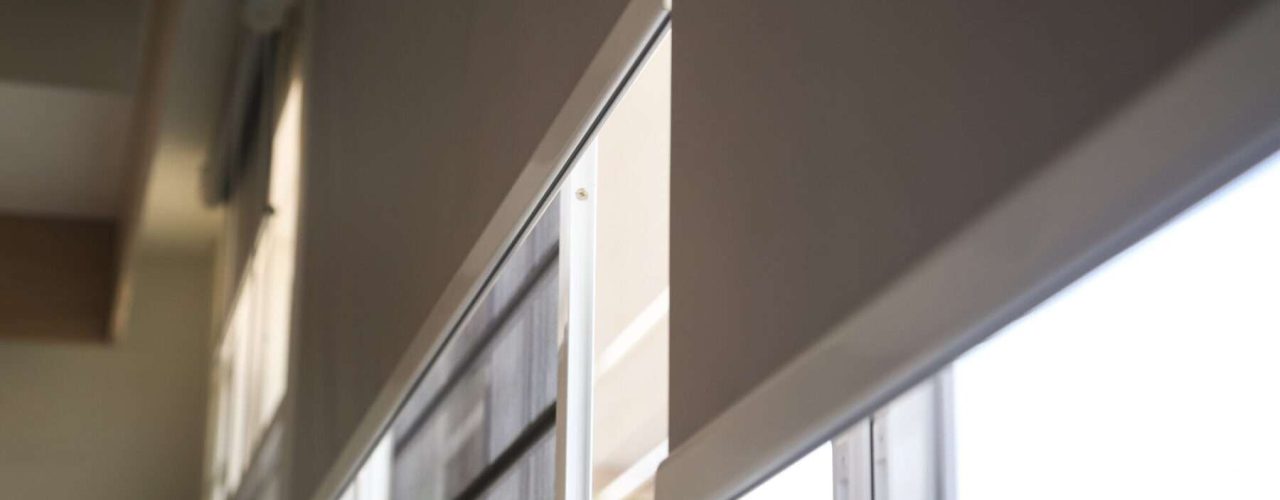 Two grey roller blinds half closed over windows with a green hedge visible through the window.