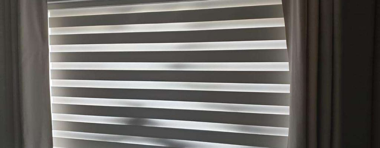 A closed day and night blind in white and grey with cream curtains drawn back at either side.