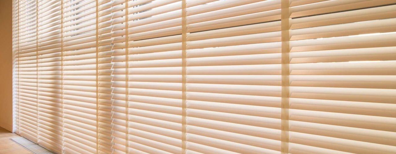 A very broad window covered in extra-wide wooden Venetian blinds in a light cream colour above a wood-effect floor.