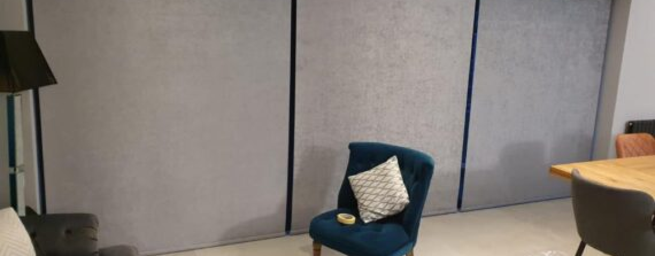 Grey motorised blinds covering a three-panel patio door, with a blue chair on a cream carpet in the foreground, a dark grey sofa to the left, and the end of a wooden table to the right.