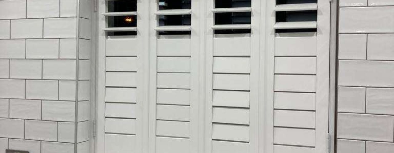 White shutters closed above a kitchen sink and white countertop, with the surrounding wall tiled in white.