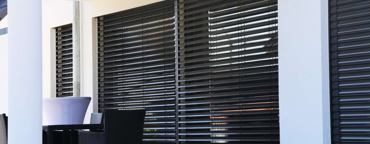 Black Venetian blinds closed over a large patio window, with a white column visible to the left alongside a black chair.