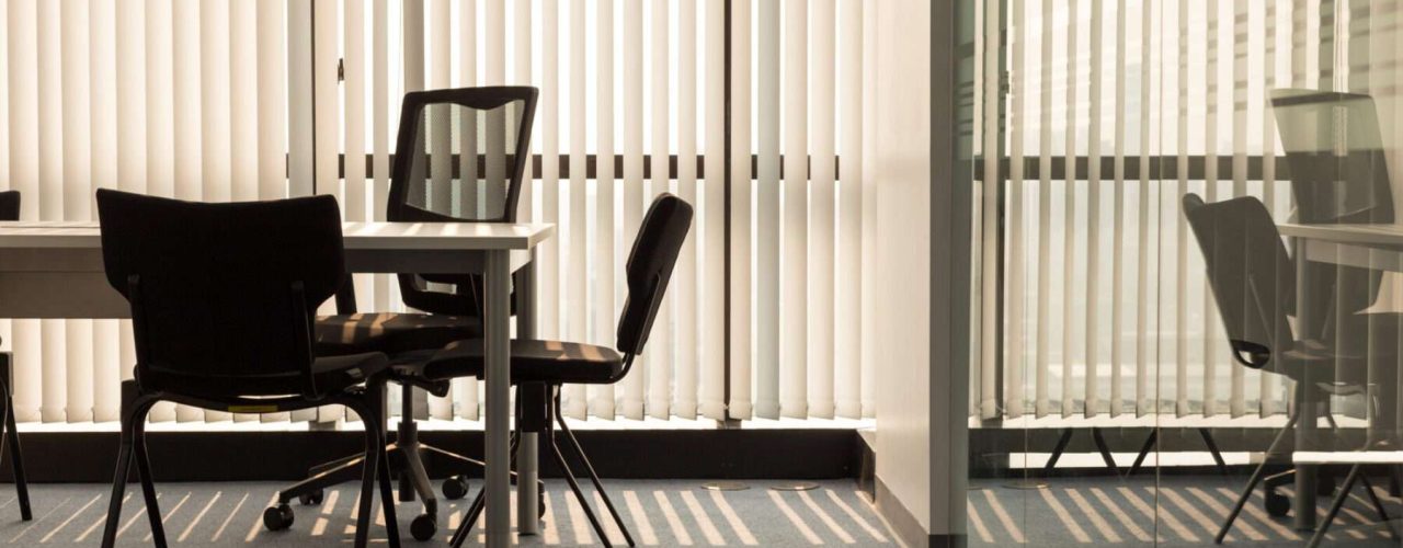 Black office chairs around a white desk, with windows covered by vertical blinds and the background, all reflected in a glass partition to the right.