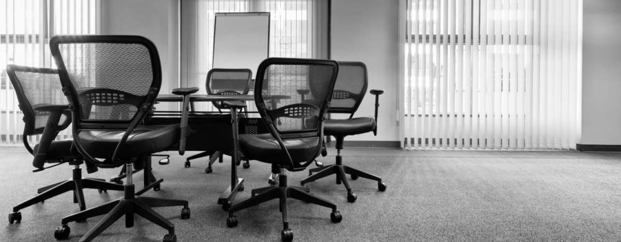 Black office chairs surrounding a small office table, with a whiteboard and white blinds decorating the windows