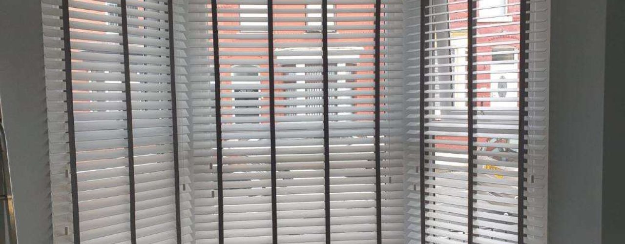 A bay window covered by white wooden Venetian blinds with black tapes, with the street outside and house opposite just visible through the slats.