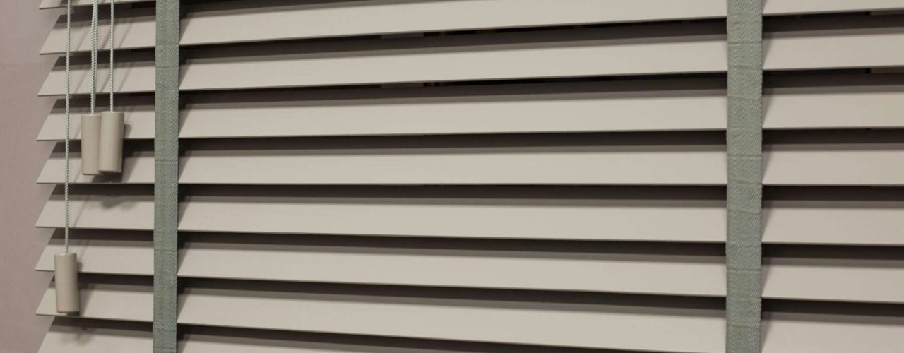 White wooden Venetian blinds partially vented with grey tapes running top to bottom at each side of the frame.